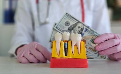 Gloved hands holding hundreds of dollars at countertop next to model dental implant