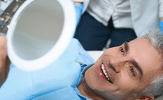 Man with gray hair reclined in dental chair smiling at reflection in handheld mirror