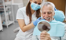 Dentist touching teeth of man with white hair and beard smiling at reflection in handheld mirror